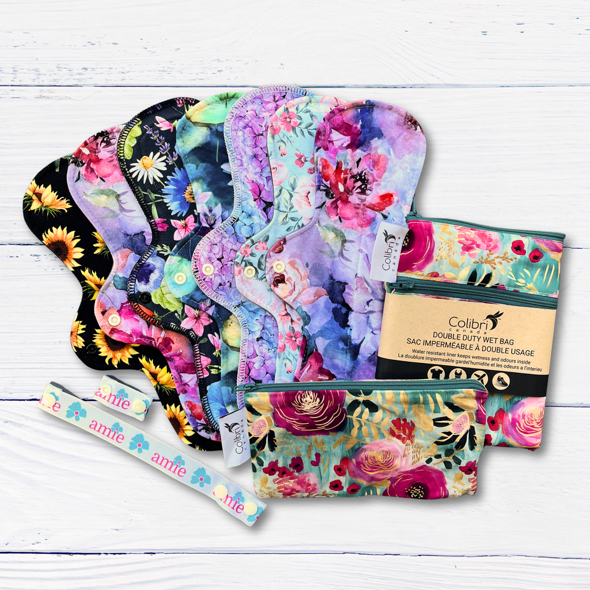 Floral-patterned long reusable pads with two colibri branded wet bags and a drying strap and wing extender on a white wooden surface
