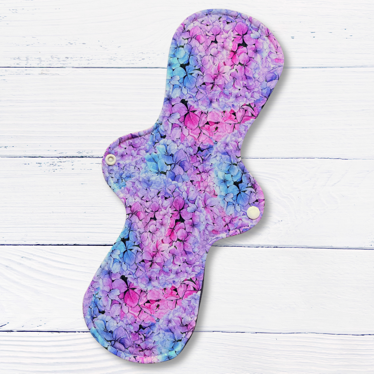 A reusable cloth sanitary pad with a vibrant pink, purple, and blue floral pattern, displayed flat on a white wood plank background.