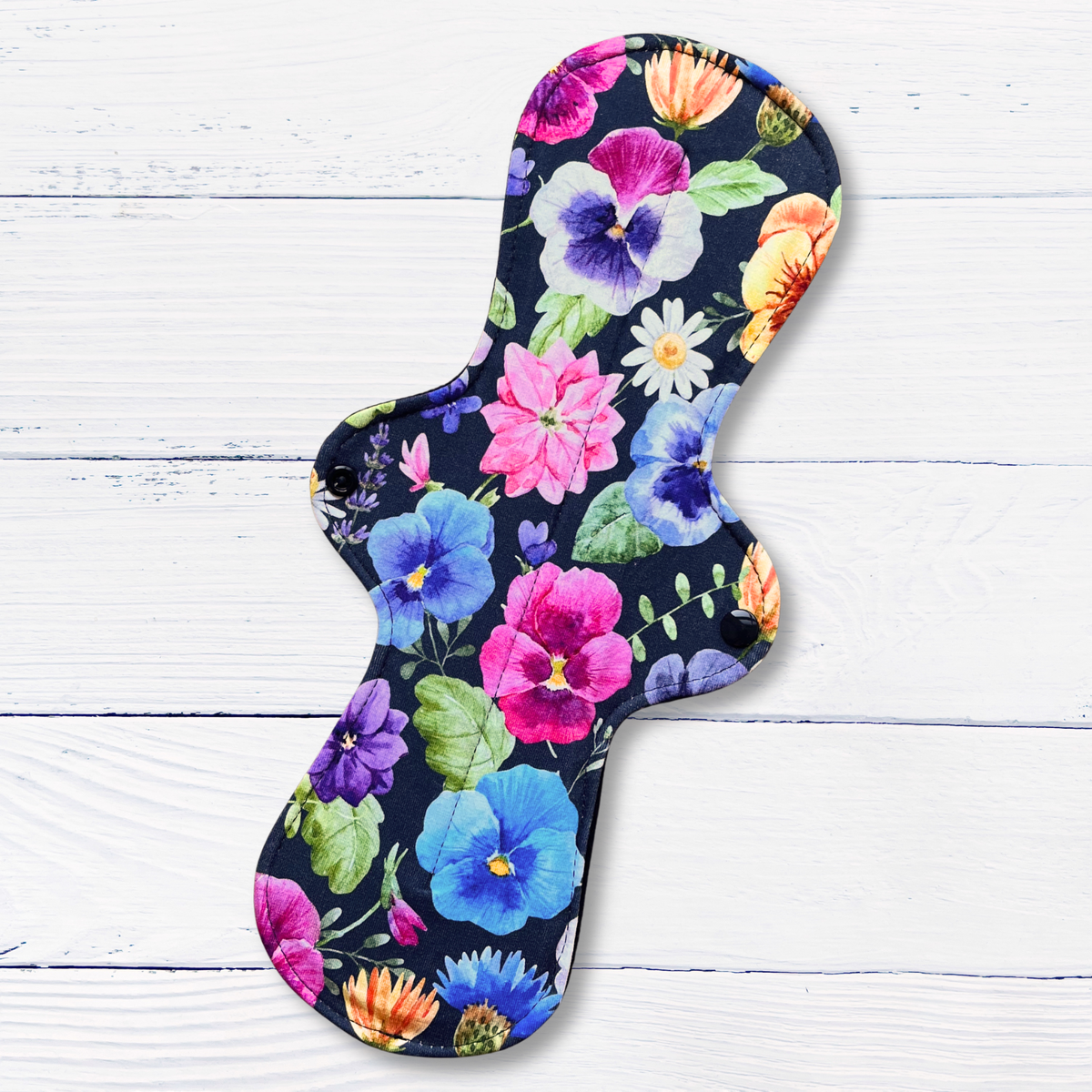 A reusable pad for periods or bladder leaks with a vibrant multicolour floral print on a dark navy background, displayed diagonally on a white wood plank surface.