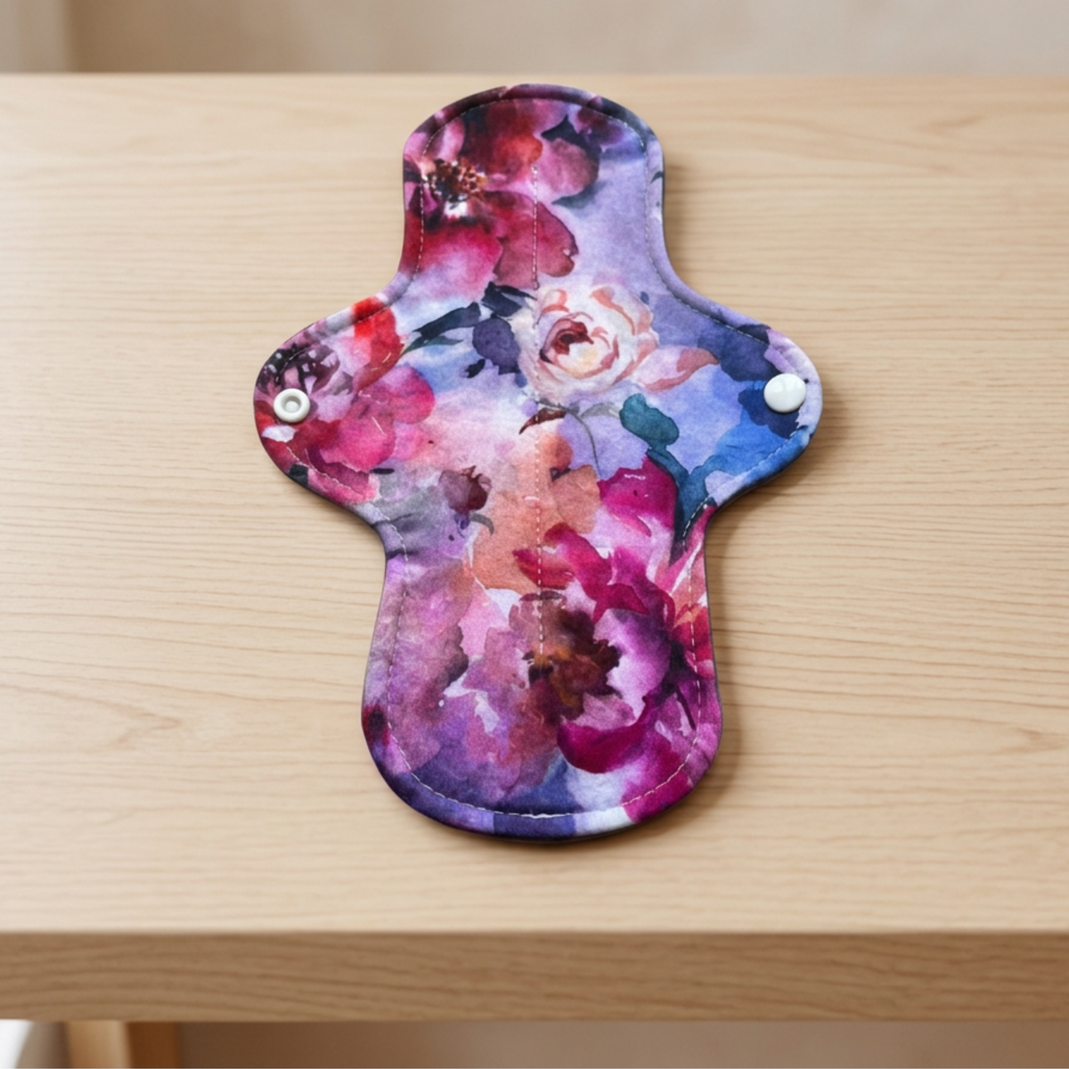 A reusable fabric sanitary pad for bladder leaks with a vibrant watercolor floral print in shades of pink, purple, and blue, resting on a light wood tabletop.