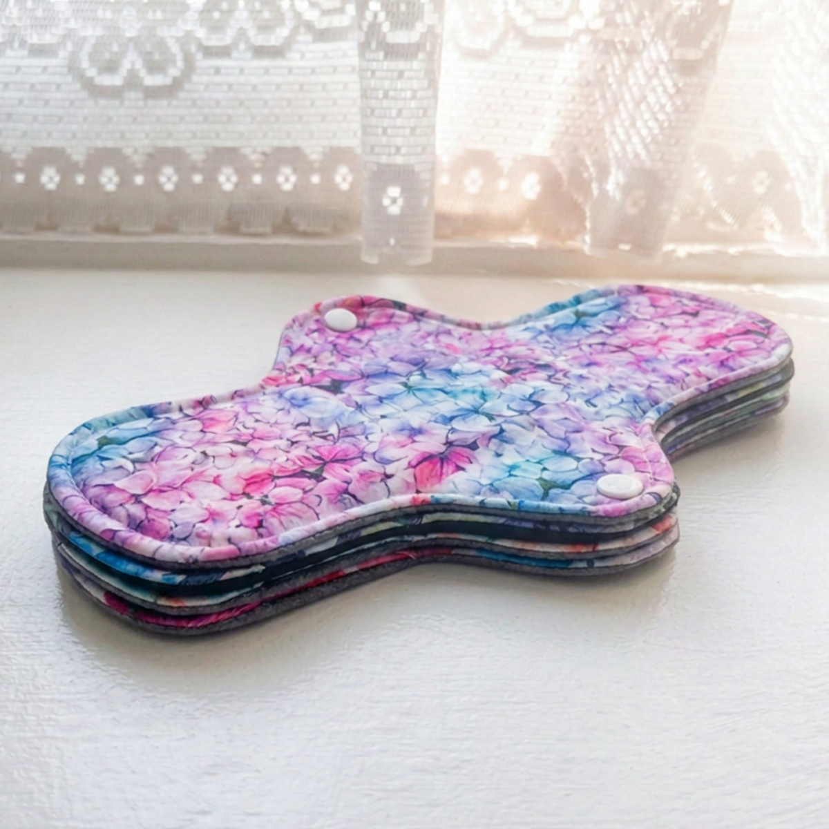 A stack of reusable sanitary pads with a colorful floral print in pink, purple, blue, and green, resting on a white surface near a lace curtain.