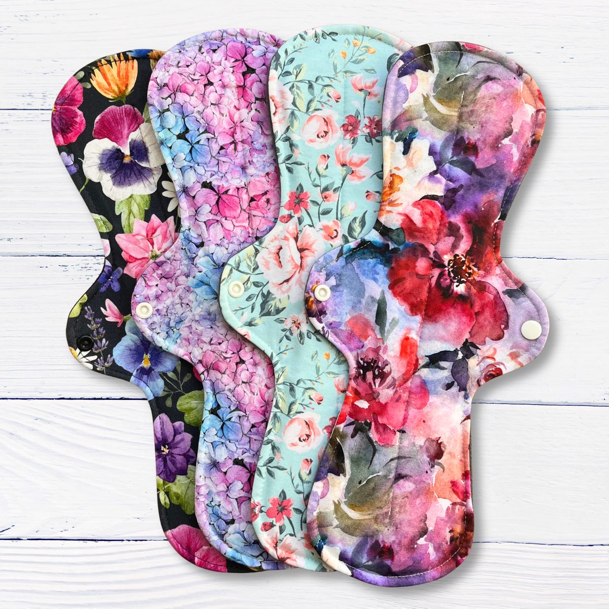 Four reusable cloth sanitary pads with floral prints are fanned out on a white wood plank background. The pads feature vibrant watercolour-style flowers in pink, purple, blue, red, and green on black, mint, and light blue backgrounds.