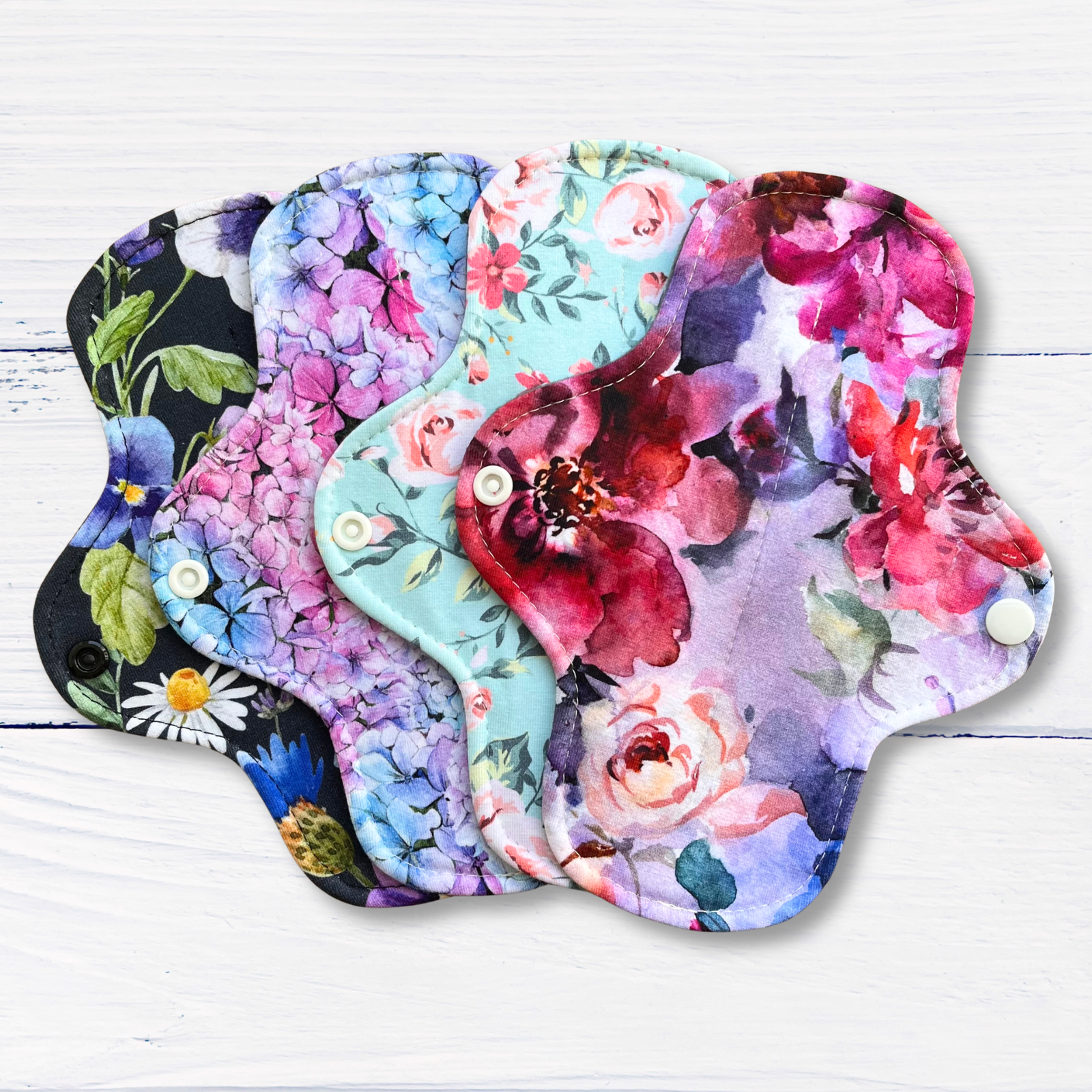 Four reusable cloth menstrual pads with floral watercolor prints are fanned out on a white wood background. The pads feature different designs including pink roses, purple hydrangeas, blue flowers, red blossoms, and a dark pattern with white snap buttons.