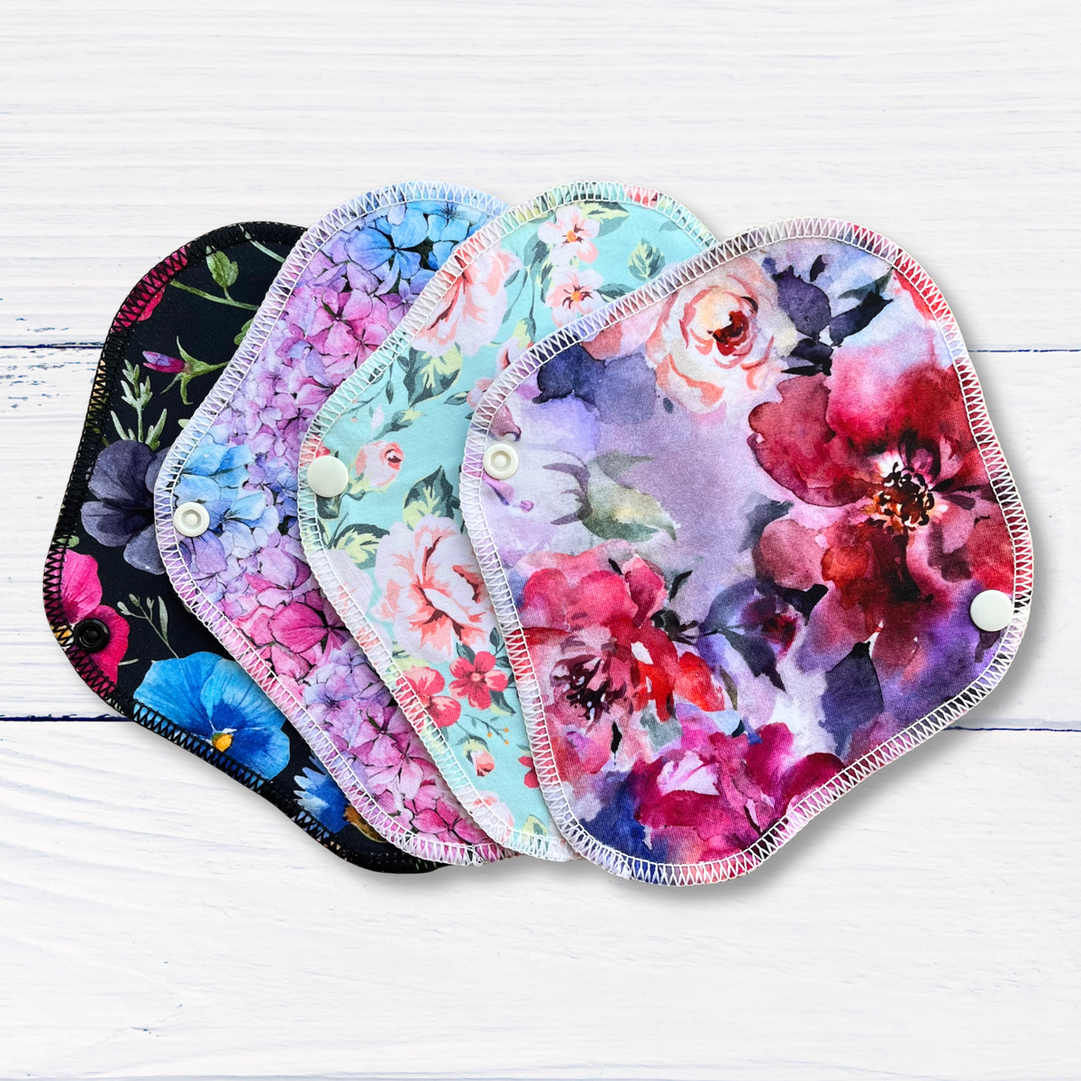 Four reusable cloth pantyliners with snap closures fanned out on a white wood background. The pads feature different floral watercolour designs in pink, purple, blue, and red tones.