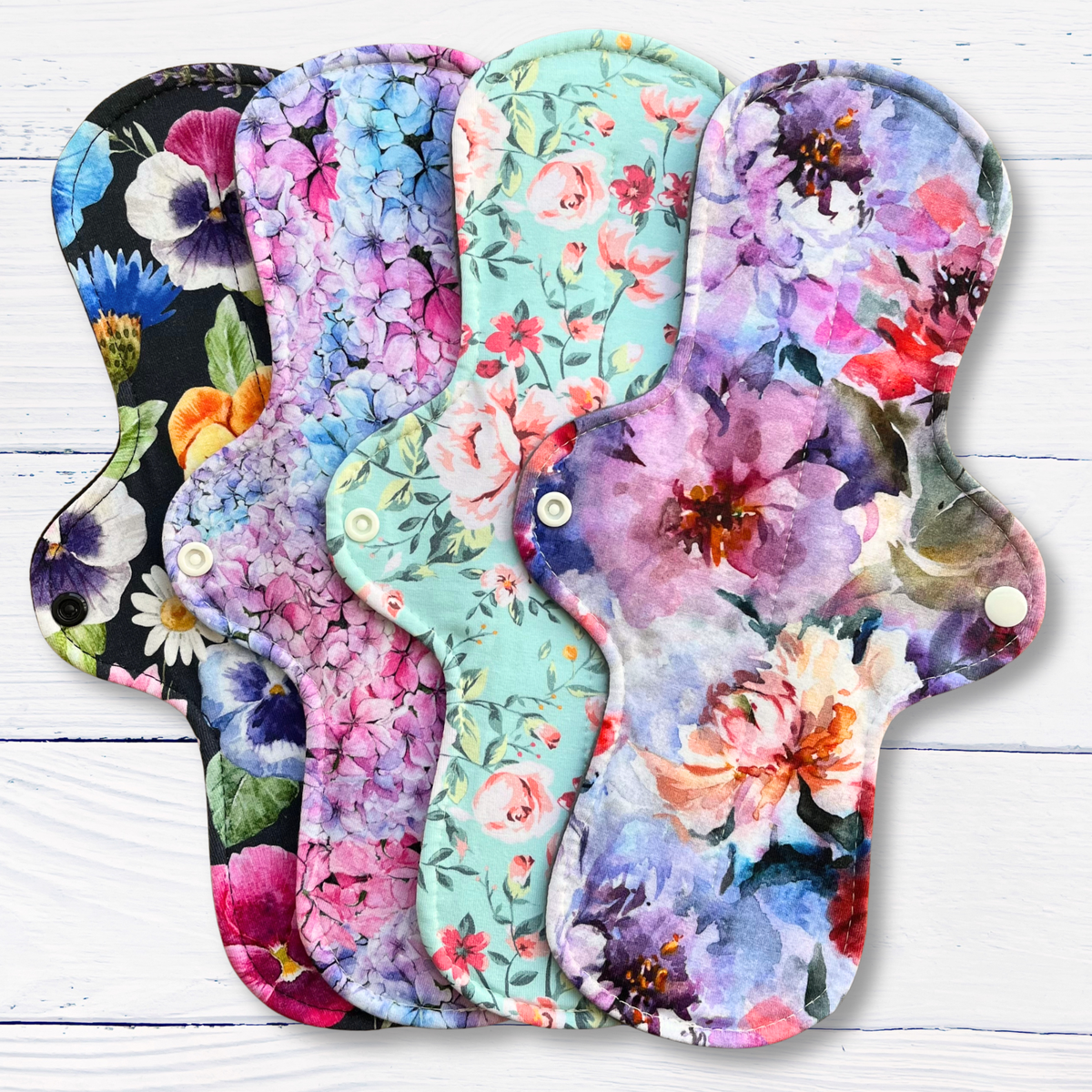 Four reusable cloth pads with different floral prints are fanned out on a white wood background. The designs include black with purple/white flowers, pink/purple hydrangeas, a mint green floral print, and purple watercolor flowers with red and orange centers.