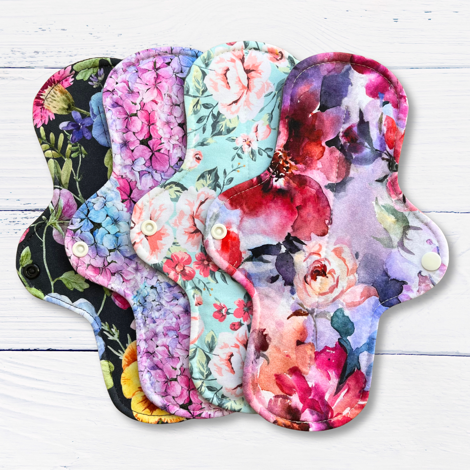 A set of reusable cloth sanitary pads with vibrant watercolour floral prints, fanned out on a white wood background.