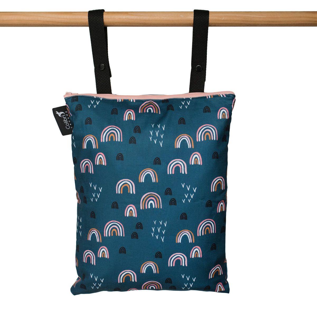 A reusable fabric tote bag with a dark teal background, featuring a repeating pattern of colorful rainbows and small raindrop shapes. The bag has black snapping straps hanging from a wooden dowel against a plain white background.