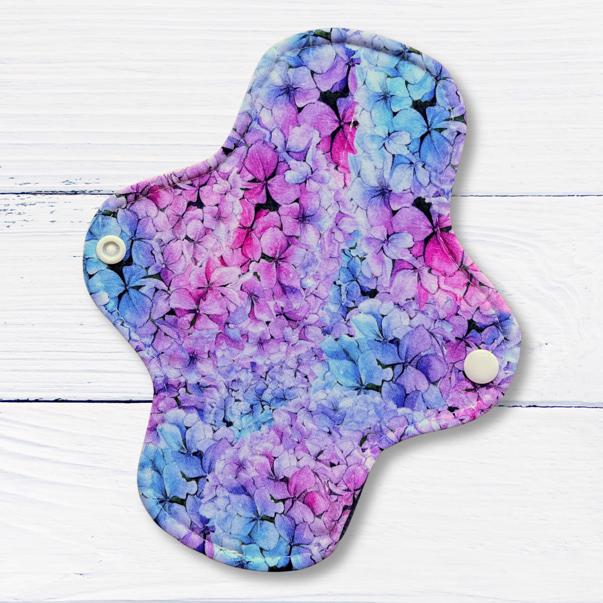 A reusable cloth sanitary pad with a vibrant purple, pink, and blue hydrangea floral print, shown on a white wood plank background.