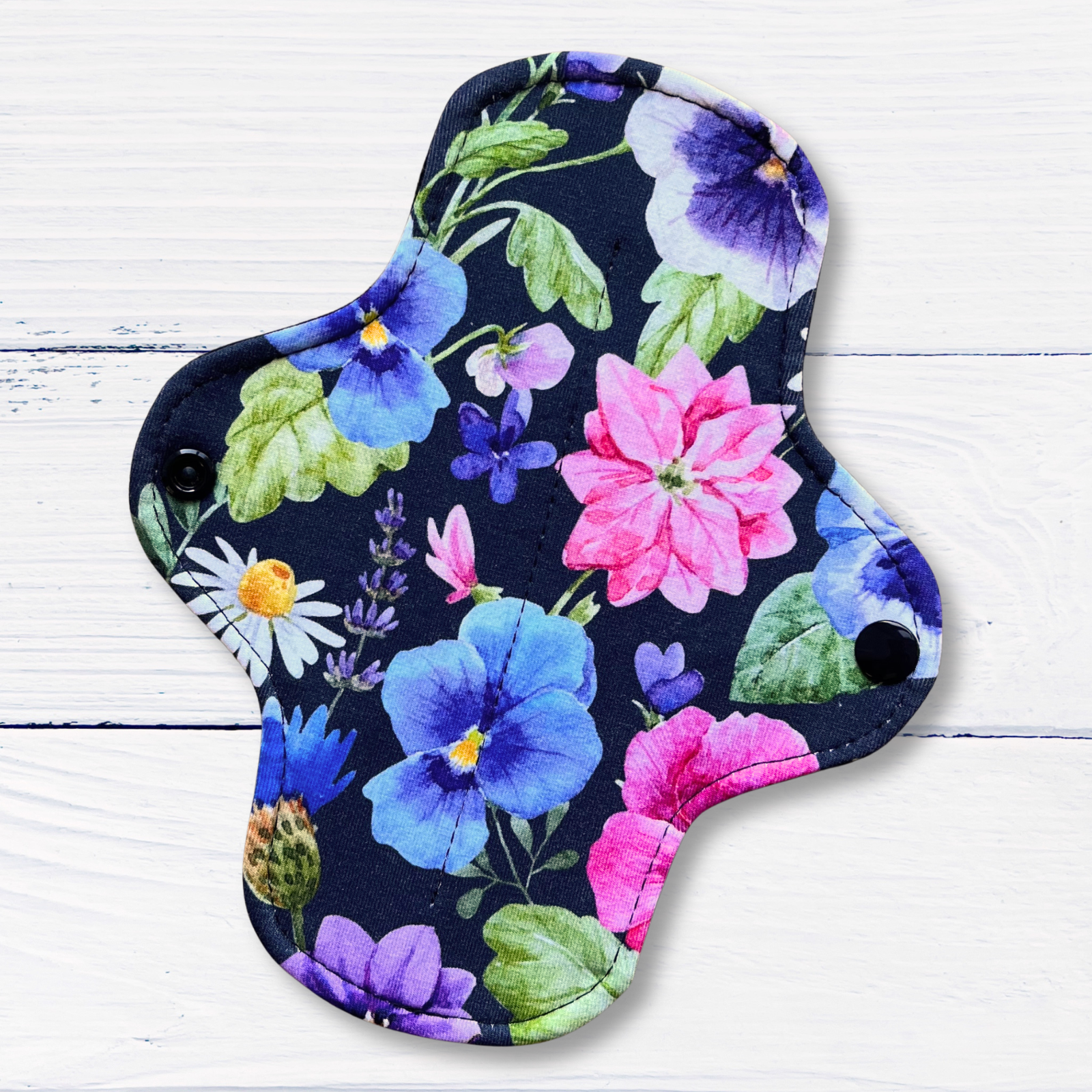 A reusable menstrual cup with a colorful floral print on a dark background, featuring pink, blue, purple, and green flowers with white snap buttons on the sides, displayed on a white wood background.