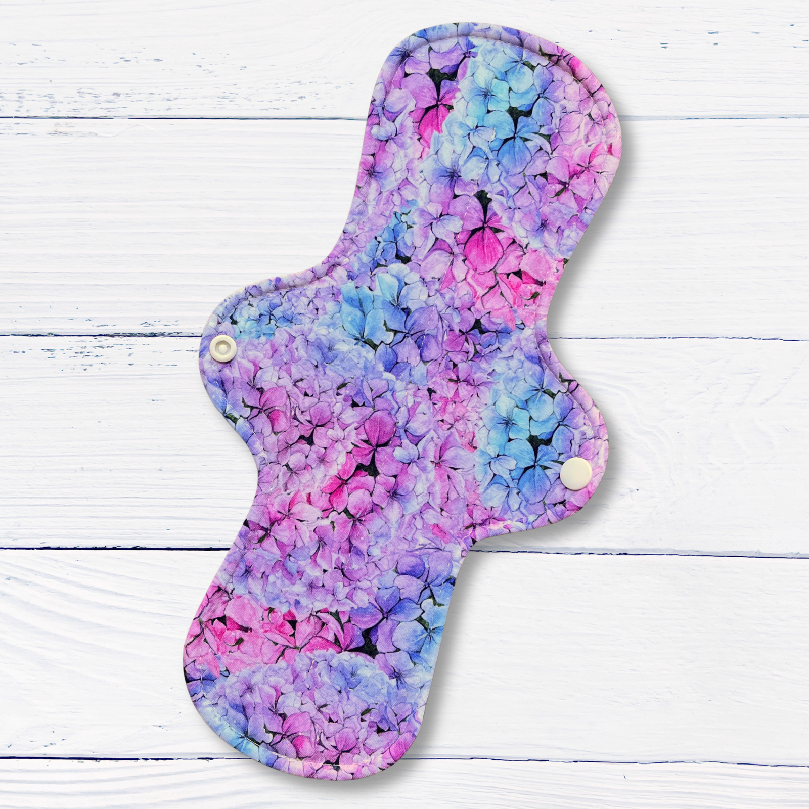 A reusable fabric pad for periods or incontinence with a vibrant purple, pink, and blue hydrangea floral print, shown on a white wood plank background.