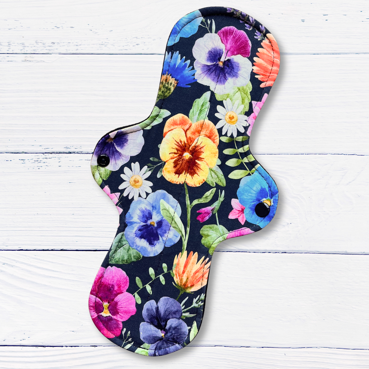 A reusable menstrual and incontinence pad with a colourful watercolour floral print on a dark navy background, displayed on a white wood plank surface.