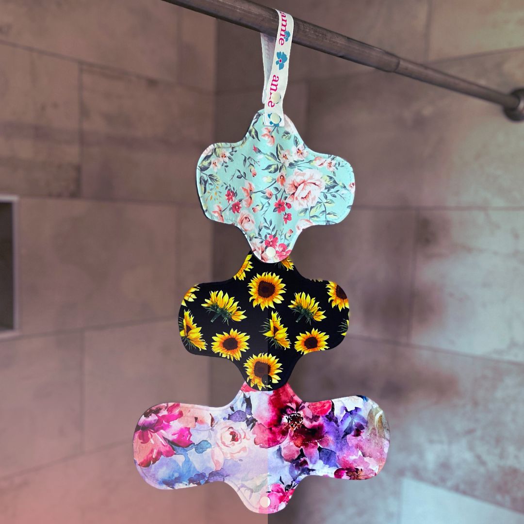 three reusable pads in floral prints snapped together in a chain hanging from a pad drying strap attached to a shower bar
