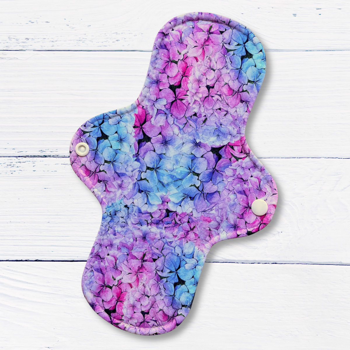 A reusable cloth pad for periods or bladder leaks with a vibrant purple, pink, and blue floral pattern, featuring two white snap buttons, displayed on a white wood plank background.
