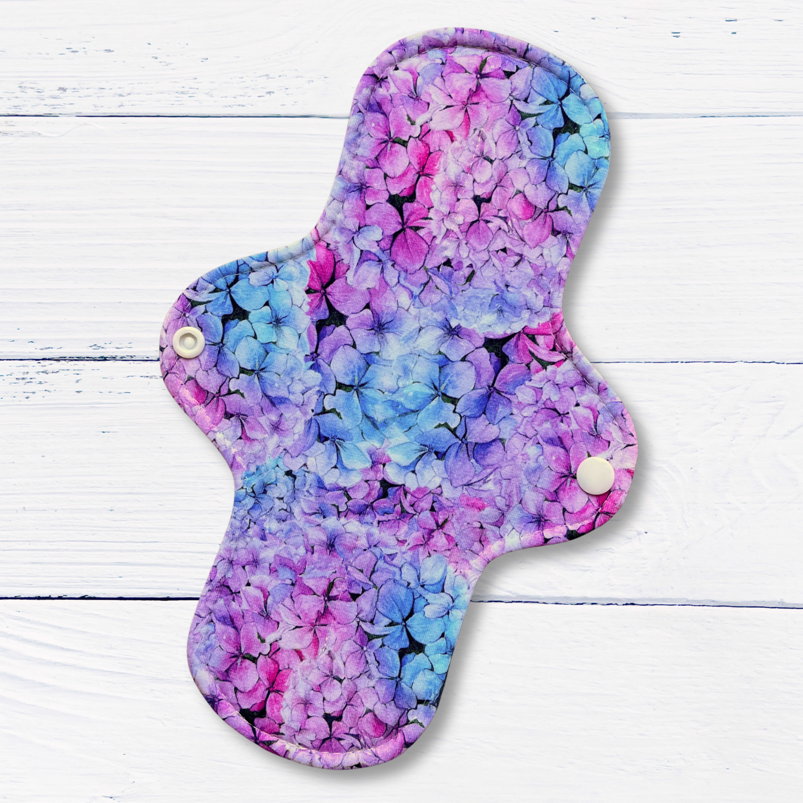 A reusable cloth pad for periods or bladder leaks with a vibrant purple, pink, and blue floral pattern, featuring two white snap buttons, displayed on a white wood plank background.