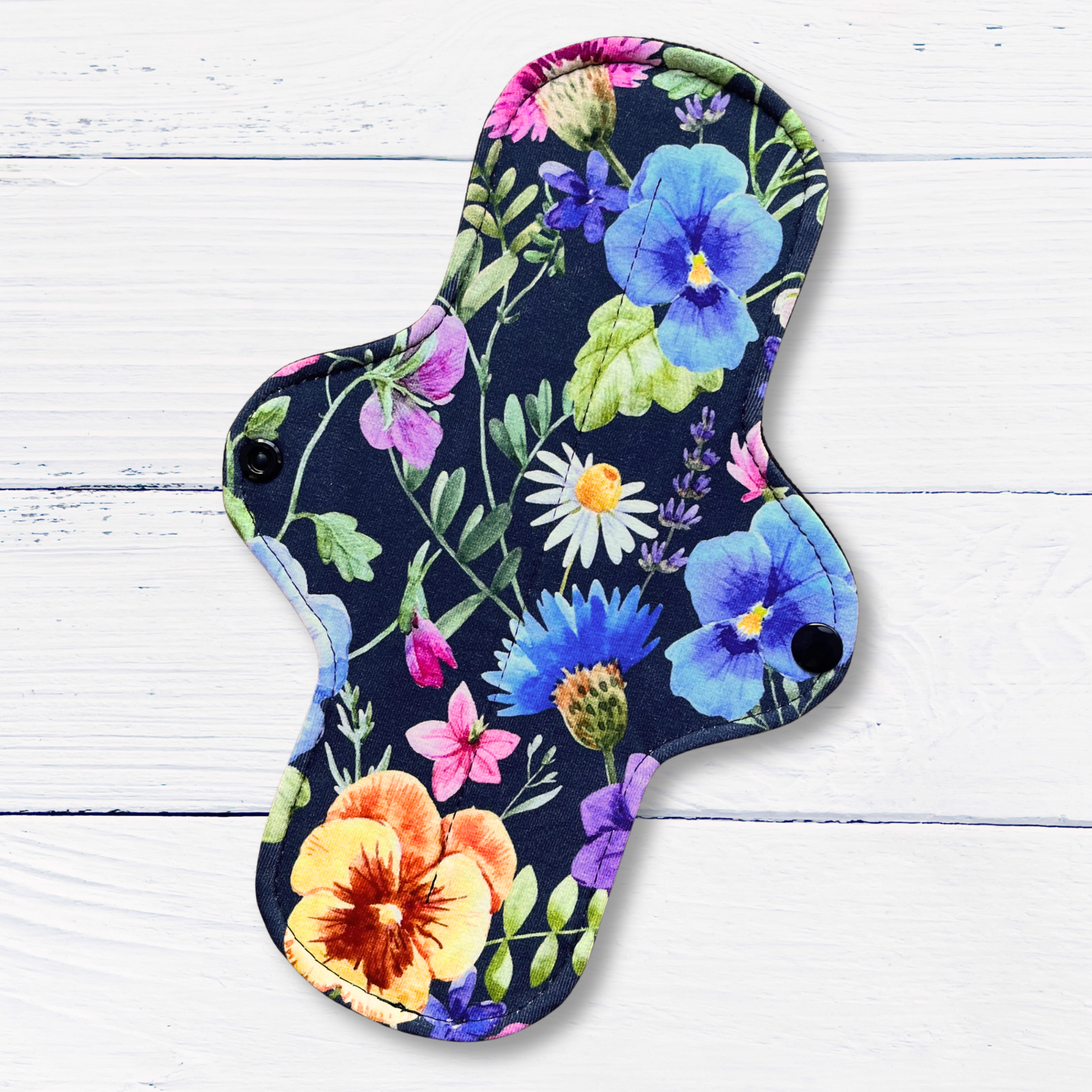 A reusable pad with a colorful floral print on a dark navy background, laid flat on a white wood plank surface.