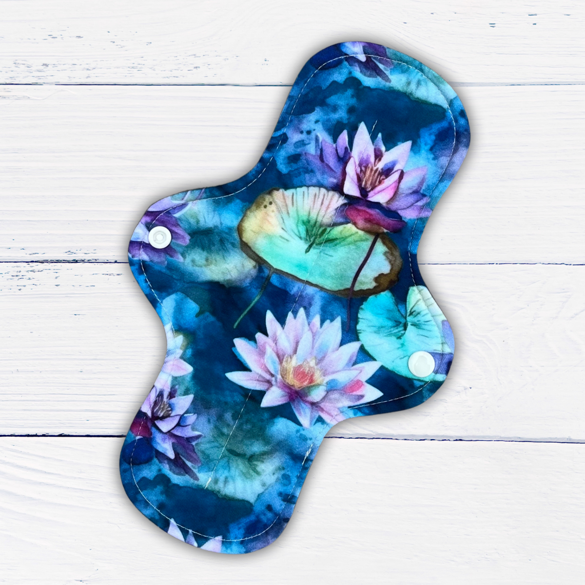 standard length moderate absorbency reusable pad in a water lily print in shades of blue green and purple on a rustic white wood background