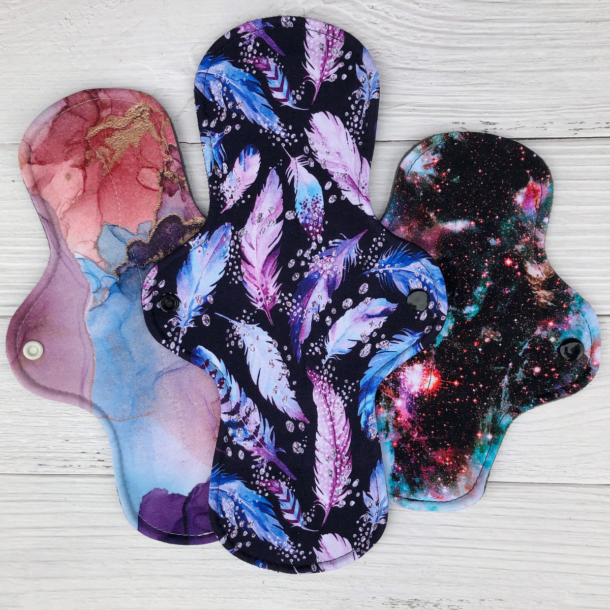 set of 3 reusable pads in prints of amtheyst purple and blue feathers and marble galaxy with black snaps on rustic wood background