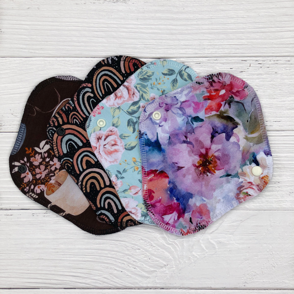 set of four reusable liners fanned out on a white wood background in the following prints - coffee cups, neutral rainbow on dark brown, mint floral and peonies