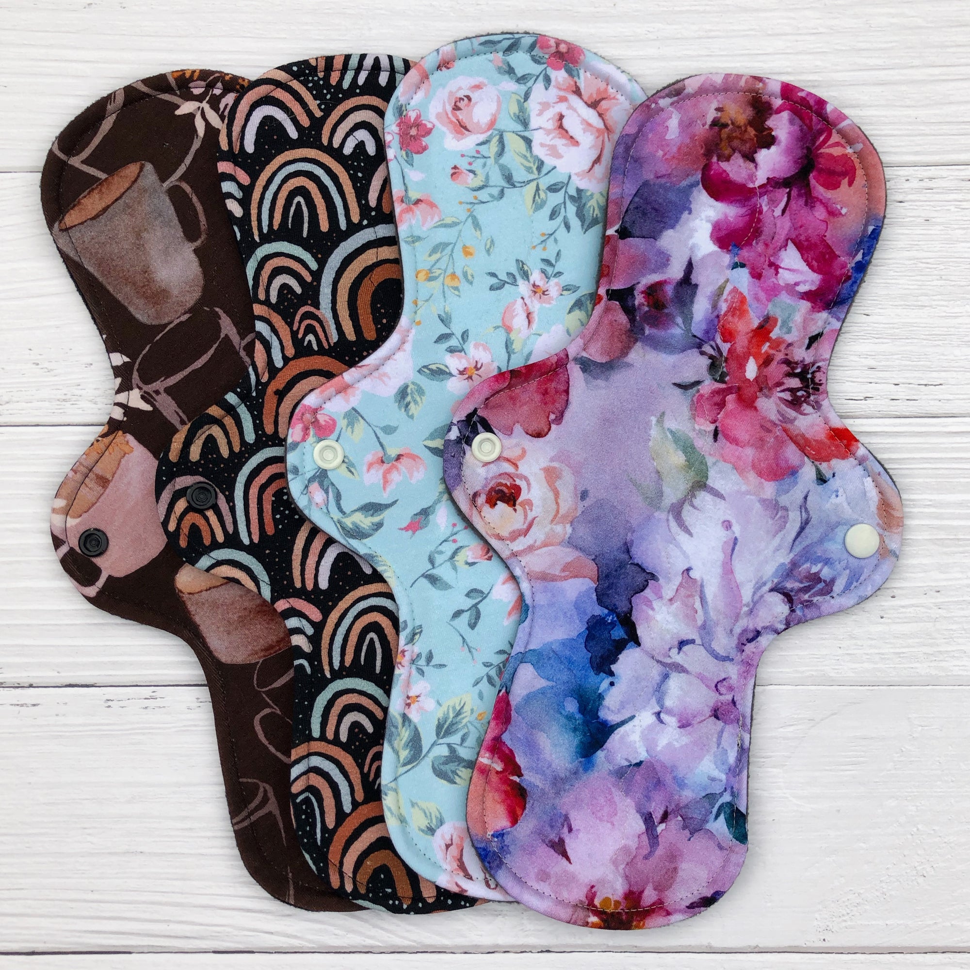 set of four reusable pads fanned out on a white wood background in the following prints - coffee cups, neutral rainbow on dark brown, mint floral and peonies